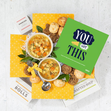 Overhead view of a yellow gift box arrangement with two bowls of soup, rolls, cookies, a container labeled “Handcrafted Soup,” a metal ladle with a yellow ribbon, and a green card that reads “You’ve Got This.”