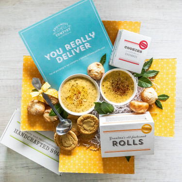Overhead view of a yellow gift box arrangement with two bowls of soup, rolls, cookies, a container labeled “Handcrafted Soup,” a metal ladle, and a blue card that reads “You Really Deliver.”