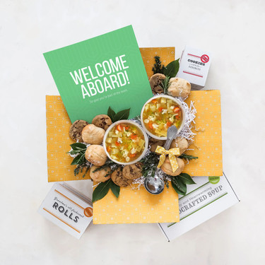 Overhead view of a yellow gift box arrangement with two bowls of soup, rolls, cookies, a container labeled “Handcrafted Soup,” a metal ladle, greenery, and a green card that reads “Welcome Aboard!”