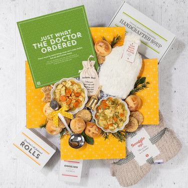 Overhead view of a yellow gift box filled with two bowls of soup, rolls, cookies, a container of handcrafted soup, a cozy throw blanket, a Warm Fuzzies warm/cool pack, and a green card that reads “Just What the Doctor Ordered.”