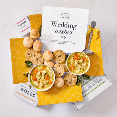 Overhead view of a yellow gift box arrangement with two bowls of soup, rolls, cookies, a container labeled “Handcrafted Soup,” a metal ladle with a yellow ribbon, and a card that reads “Wedding Wishes.”