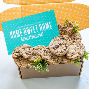 Open yellow gift box filled with assorted cookies and greenery, with a teal card that reads “Home Sweet Home Congratulations!”