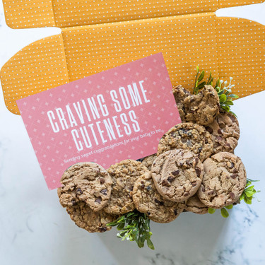 Open yellow gift box filled with assorted cookies and greenery, with a pink card that reads “Craving Some Cuteness.”
