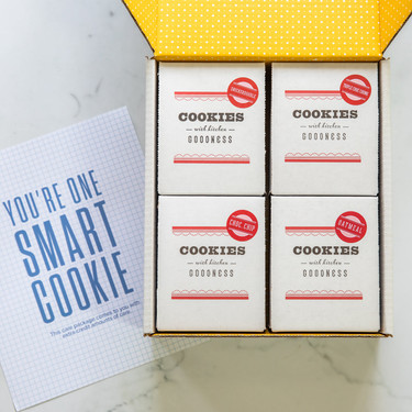 Open yellow gift box containing four cookie boxes next to a card that reads “You’re One Smart Cookie.”