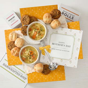 Overhead view of a yellow gift box arrangement with two bowls of soup, rolls, cookies, a container labeled “Handcrafted Soup,” a metal ladle with a yellow ribbon, and a card that reads “Happy Mother’s Day.”