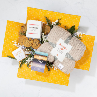 Overhead view of a yellow gift box containing a cozy knit throw blanket, a box of cookies, assorted cookies, tea sachets, a jar of honey with a honey dipper, a healing-after-loss book, and greenery.