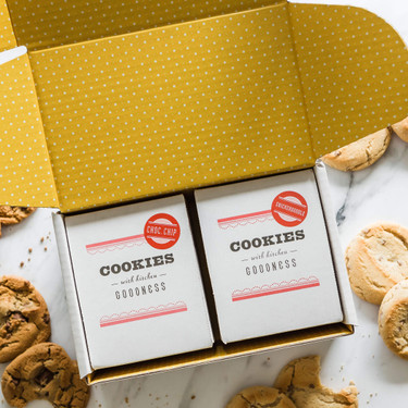 Open yellow box with white polka dots containing two packages labeled “Cookies” surrounded by assorted cookies on a marble surface.