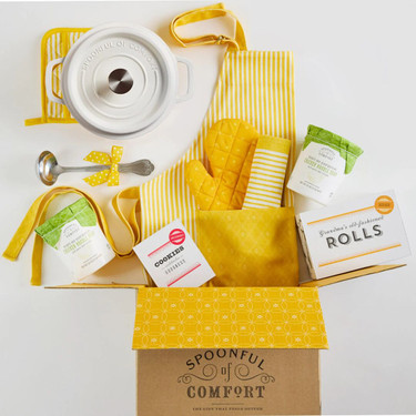 Open Spoonful of Comfort gift box filled with yellow kitchen items including a Dutch oven with lid, utensils, a towel, apron, and oven mitt, along with packaged soup containers, cookies, and rolls arranged inside and around the box.
