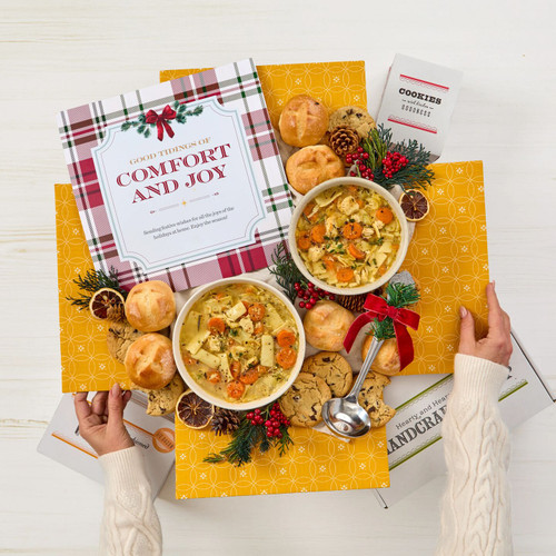 Overhead view of hands holding a yellow gift box arrangement with two bowls of soup, rolls, cookies, a container labeled “Handcrafted Soup,” a metal ladle with a yellow ribbon, holiday greenery, and a card that reads “Comfort and Joy.”