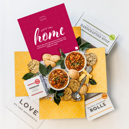 Unboxed College Care Package with an insert card with text that reads Feels Like Home. Displayed with two bowls of chicken noodle soup, chocolate chip cookies and dinner rolls.