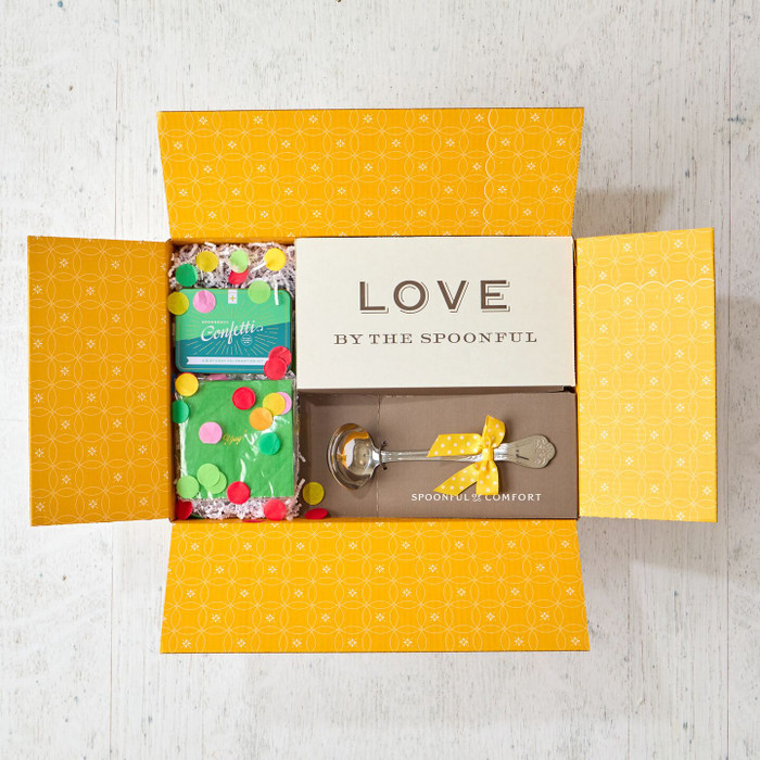 Open yellow gift box displaying a card that reads “Love by the Spoonful,” a metal ladle with a yellow ribbon, and two confetti poppers with colorful confetti packaging.