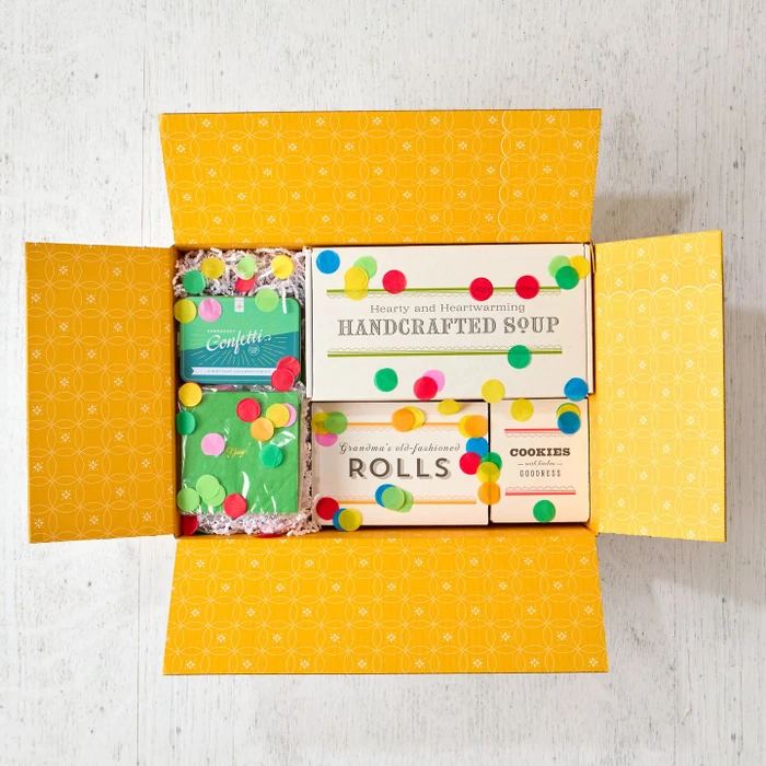Open yellow gift box filled with packages labeled “Handcrafted Soup,” “Rolls,” and “Cookies,” alongside two confetti poppers and scattered colorful confetti.