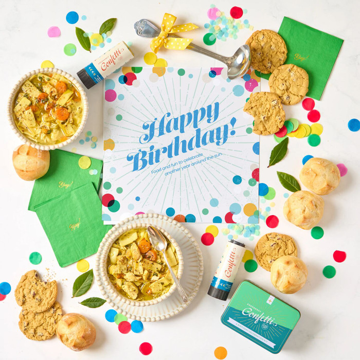 Overhead view of a birthday-themed gift arrangement with two bowls of soup, rolls, cookies, a container labeled “Handcrafted Soup,” a metal ladle with a yellow ribbon, green birthday napkins, confetti poppers, colorful confetti, and a card that reads “Happy Birthday!”