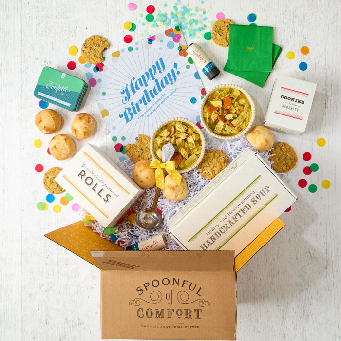 Open Spoonful of Comfort gift box with a birthday-themed arrangement including two bowls of soup, rolls, cookies, colorful confetti, green napkins, confetti poppers, packaged items, and a card that reads “Happy Birthday!”