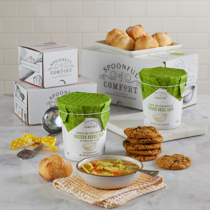 Soup gift set on a counter with a bowl of soup, rolls, cookies, two containers of soup with green lids, stacked Spoonful of Comfort boxes, a metal ladle with a yellow ribbon, and a folded towel.