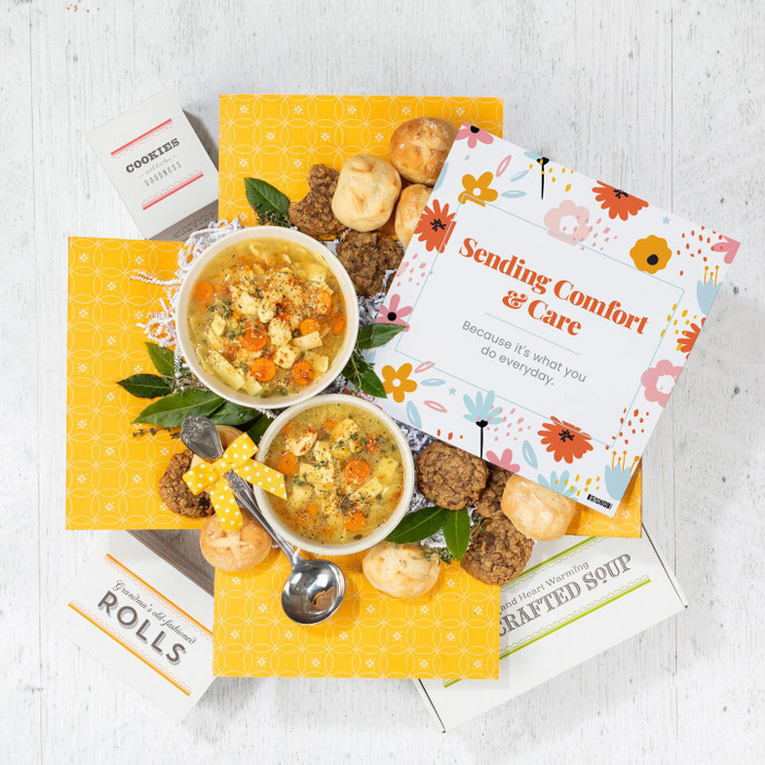 Overhead view of a yellow gift box arrangement with two bowls of soup, rolls, cookies, a container labeled “Handcrafted Soup,” a metal ladle with a yellow ribbon, and a floral card that reads “Sending Comfort & Care.”