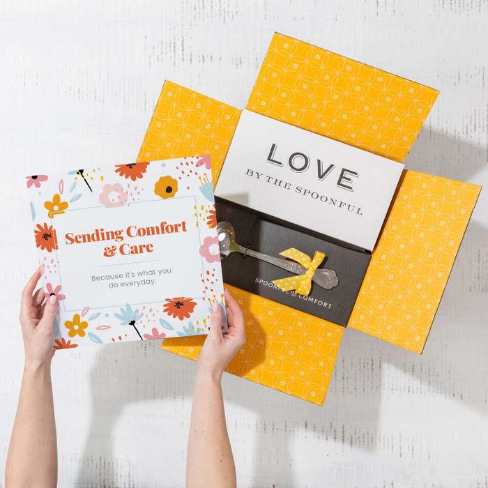 Overhead view of hands holding a floral card that reads “Sending Comfort & Care” next to an open yellow gift box with a card that reads “Love by the Spoonful” and a metal ladle with a yellow ribbon inside.