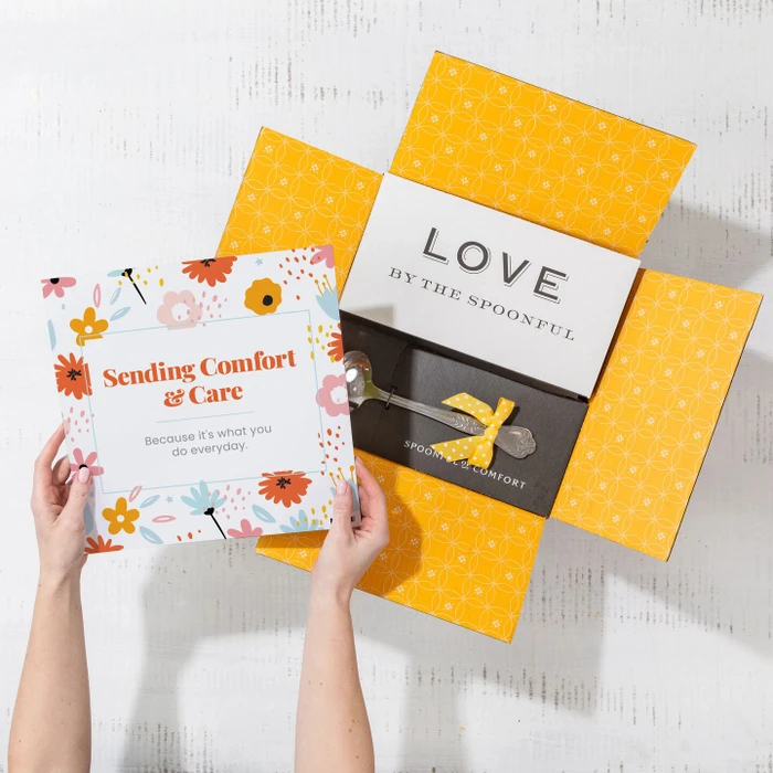 Overhead view of hands holding a floral card that reads “Sending Comfort & Care” next to an open yellow gift box with a card that reads “Love by the Spoonful” and a metal ladle with a yellow ribbon inside.
