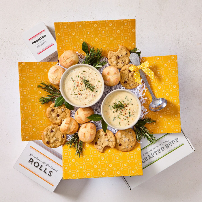 Overhead view of a yellow gift box arrangement with two bowls of creamy soup, rolls, cookies, a container labeled “Handcrafted Soup,” and a metal ladle with a yellow ribbon, arranged with greenery.