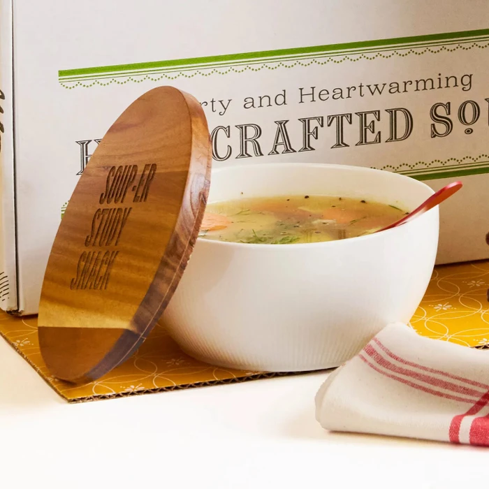 Close-up of a bowl of soup with a wooden lid resting beside it, set in front of a box labeled “Handcrafted Soup.”