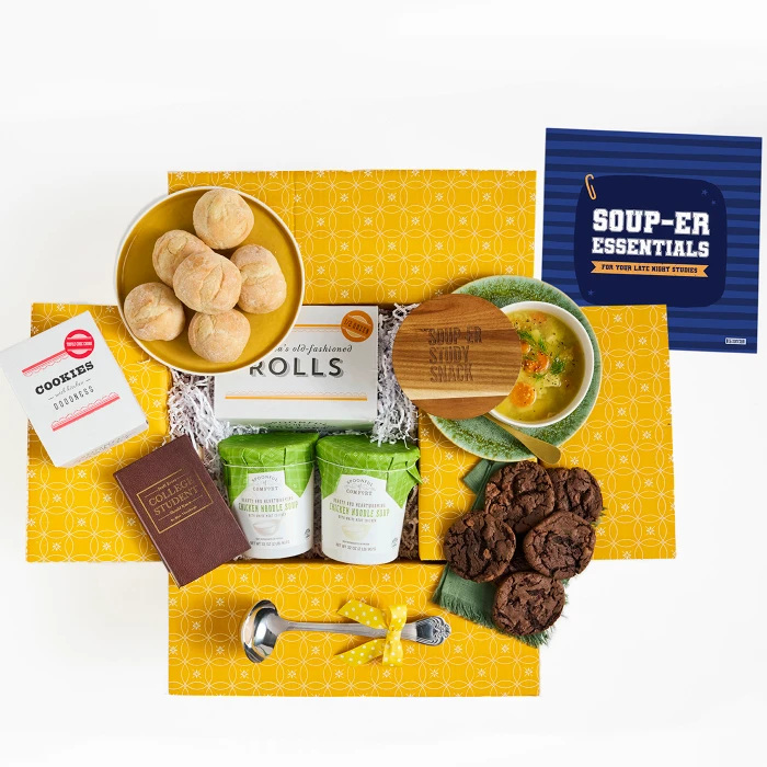 Overhead view of a yellow gift box arrangement with a bowl of soup, rolls, cookies, two containers of soup, a metal ladle, a brown book labeled “College Essentials,” and a blue card that reads “Soup-er Essentials.”