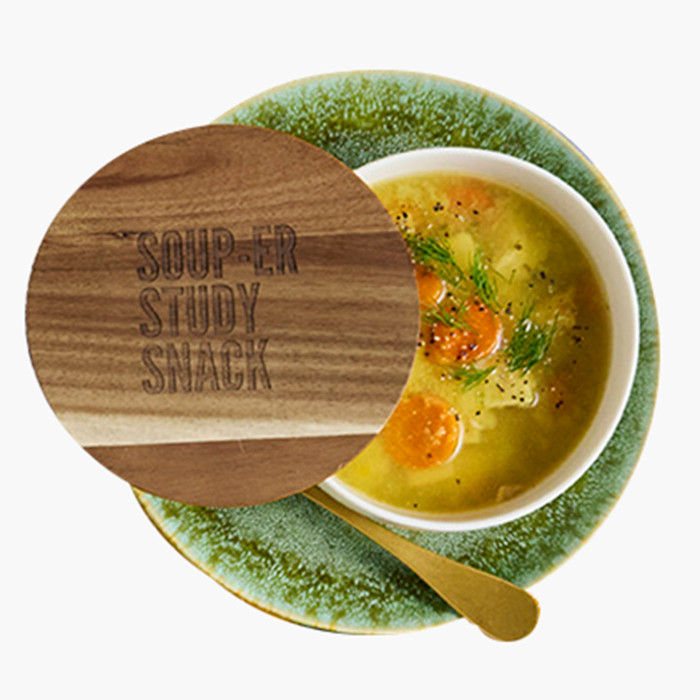 Close-up of a bowl of soup on a green plate with a wooden lid engraved “Soup-er Study Snack,” partially covering the bowl.
