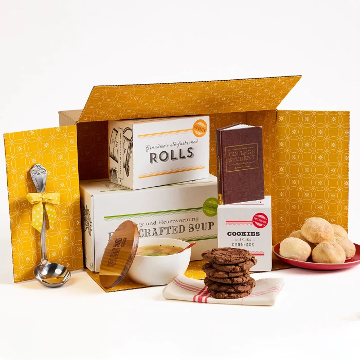 Open yellow gift box containing packaged rolls, a container labeled “Handcrafted Soup,” a box of cookies, a stack of cookies, rolls on a plate, a metal ladle with a yellow ribbon, and a small book.