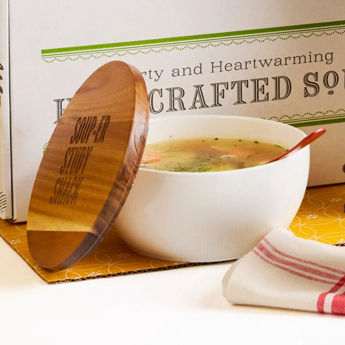 Close-up of a bowl of soup with a wooden lid resting beside it, set in front of a box labeled “Handcrafted Soup.”