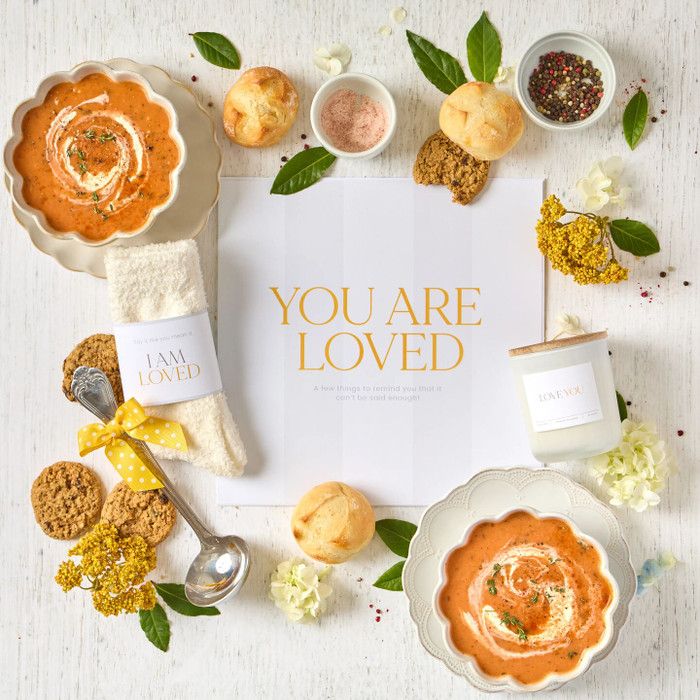 Overhead view of a comforting gift arrangement with two bowls of tomato soup, rolls, cookies, a candle, cozy socks labeled “I Am Loved,” a metal ladle with a yellow ribbon, greenery, and an open card that reads “You Are Loved.”