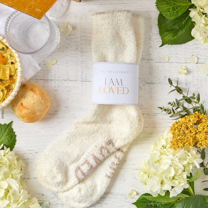 Close-up of a pair of cozy cream-colored socks labeled “I Am Loved,” surrounded by flowers, greenery, and nearby food items.