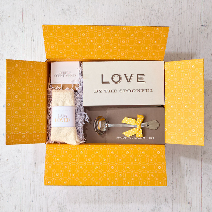 Open yellow gift box displaying a card that reads “Love by the Spoonful,” a pair of cozy socks labeled “I Am Loved,” and a metal ladle with a yellow ribbon inside.