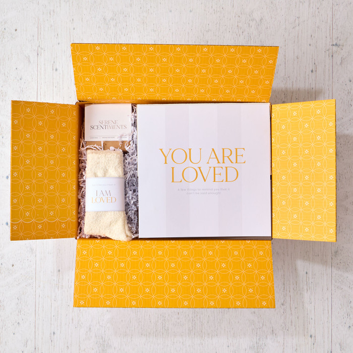 Open yellow gift box displaying a card that reads “You Are Loved” alongside a pair of cozy socks labeled “I Am Loved.”
