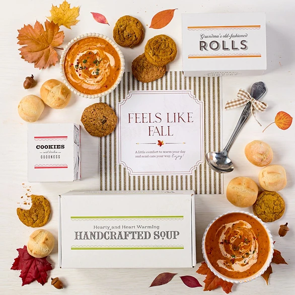 Overhead view of a fall-themed arrangement with two bowls of soup, rolls, cookies, a box labeled “Handcrafted Soup,” a box of rolls, a metal ladle with a yellow ribbon, autumn leaves, and a card that reads “Feels Like Fall.”