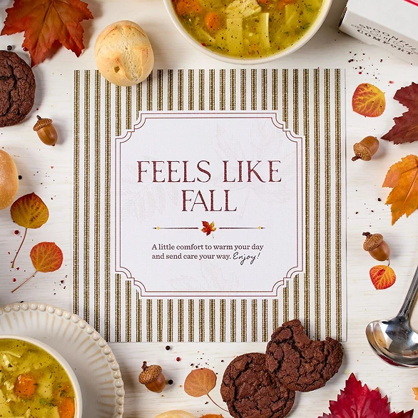 Overhead view of a fall-themed arrangement with two bowls of soup, rolls, cookies, a box labeled “Handcrafted Soup,” a box of rolls, a metal ladle with a yellow ribbon, autumn leaves, and a card that reads “Feels Like Fall.”