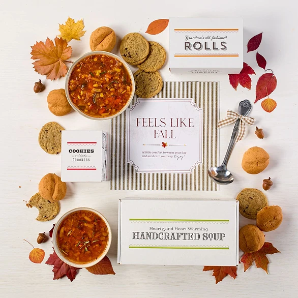 Overhead view of a fall-themed gift arrangement with two bowls of soup, rolls, cookies, a container labeled “Handcrafted Soup,” a metal ladle with a yellow ribbon, autumn leaves, and a card that reads “Feels Like Fall.”