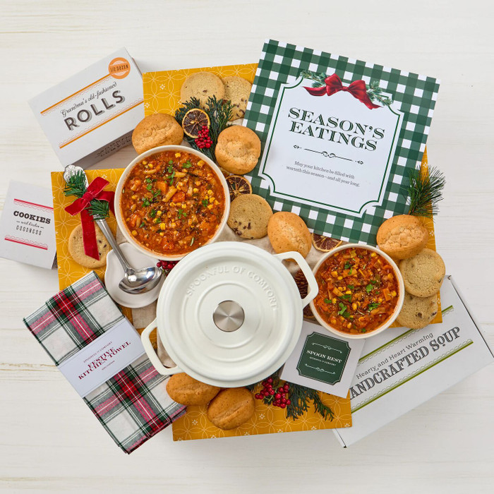 Overhead view of a holiday gift arrangement with two bowls of soup, rolls, cookies, a container labeled “Handcrafted Soup,” a white Dutch oven with lid, a metal ladle with a red ribbon, a plaid kitchen towel, a spoon rest, festive greenery, and a card that reads “Season’s Eatings.”