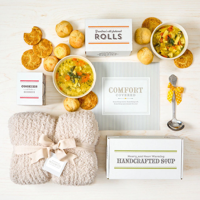 Overhead view of a comfort-themed gift arrangement with two bowls of soup, rolls, cookies, a box labeled “Handcrafted Soup,” a box of rolls, a box of cookies, a metal ladle with a yellow ribbon, a cozy knit blanket, and a card that reads “Comfort Covered.”