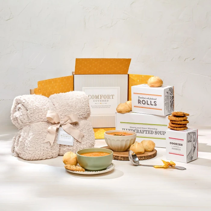 Comfort-themed gift set displayed with a cozy knit blanket, two bowls of soup, rolls, cookies, boxes labeled “Handcrafted Soup,” “Rolls,” and “Cookies,” a metal ladle with a yellow ribbon, and a card that reads “Comfort Covered.”