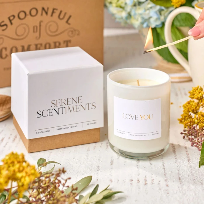 Close-up of a “Love You” candle being lit next to its box labeled “Serene Scents,” with dried flowers and a Spoonful of Comfort box in the background.