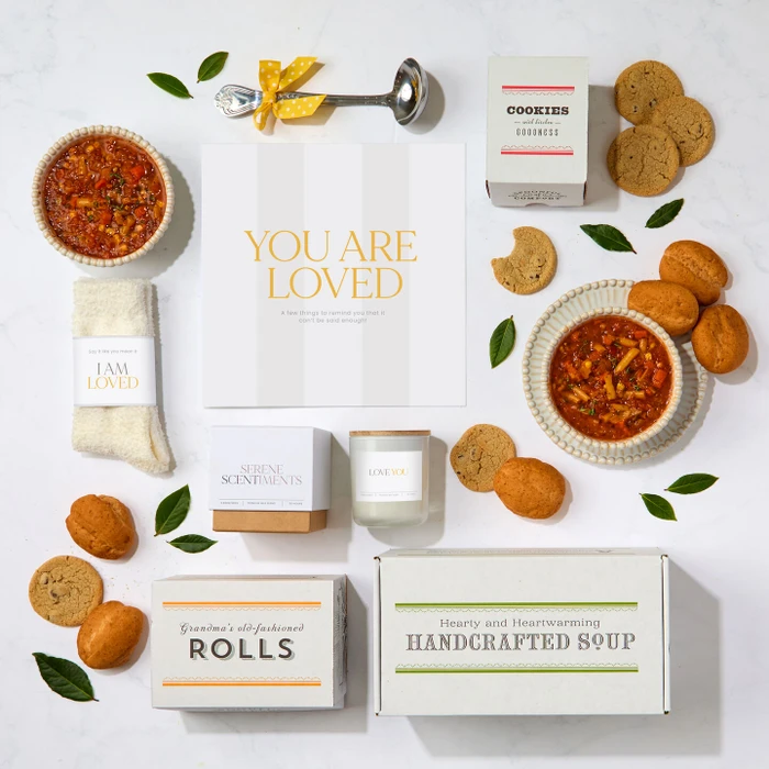 Overhead view of a comforting gift arrangement with two bowls of soup, rolls, cookies, a candle, cozy socks labeled “I Am Loved,” a box labeled “Handcrafted Soup,” a box of rolls, a box of cookies, a metal ladle with a yellow ribbon, greenery, and a card that reads “You Are Loved.”