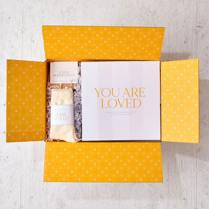 Open yellow gift box displaying a card that reads “Love by the Spoonful,” a pair of cozy socks labeled “I Am Loved,” and a metal ladle with a yellow ribbon inside.