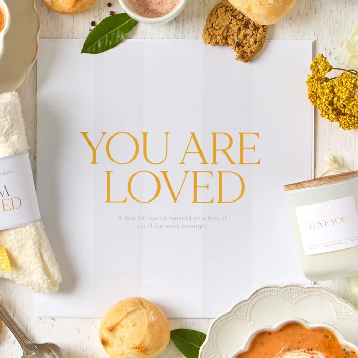 Close-up of an open card that reads “You Are Loved,” surrounded by soup, rolls, cookies, a candle, cozy socks, and greenery.