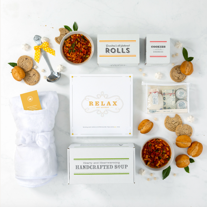 Overhead view of a relaxation-themed gift arrangement with two bowls of soup, rolls, cookies, a box labeled “Handcrafted Soup,” a robe, self-care items in a toiletry bag, a metal ladle, and a card that reads “Relax.”