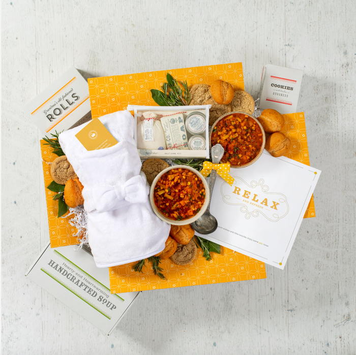 Overhead view of a yellow gift box arrangement with two bowls of soup, rolls, cookies, a container labeled “Handcrafted Soup,” a robe, self-care items in a toiletry bag, and a card that reads “Time to Relax.”