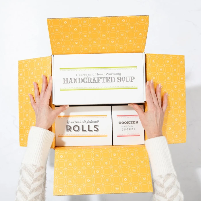 Overhead view of hands opening a yellow gift box containing packages labeled “Handcrafted Soup,” “Rolls,” and “Cookies.”
