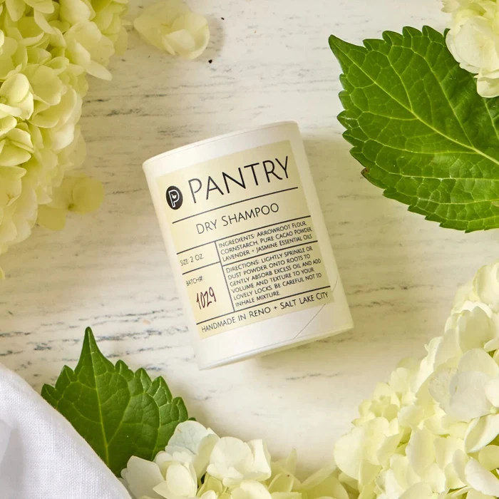 Close-up of a Pantry brand dry shampoo container surrounded by white flowers and green leaves on a light surface.