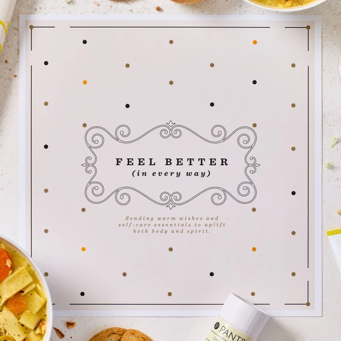 Close-up of a card that reads “Feel Better (in every way),” with decorative accents and parts of a soup and self-care gift arrangement visible around it.