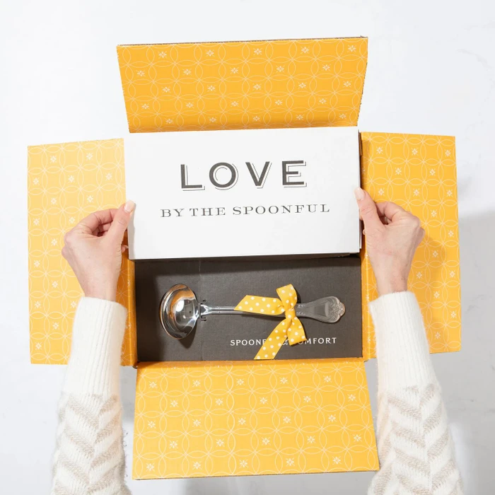 Open yellow gift box displaying a card that reads “Love by the Spoonful” and a metal ladle with a yellow ribbon inside.