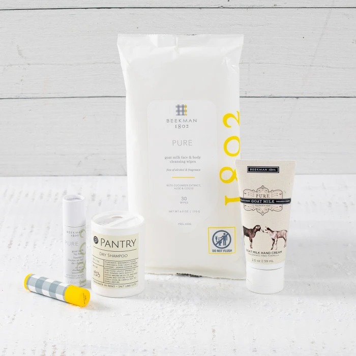 Close-up of self-care items including a large bag labeled “Cozy,” a candle, hand cream, lip balm, and a spray bottle arranged on a light surface.