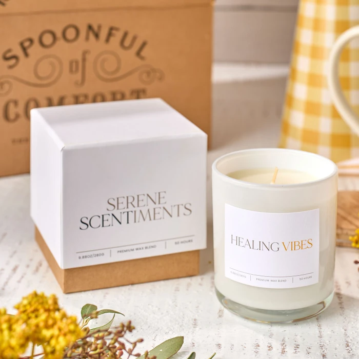 Close-up of a “Healing Vibes” candle next to its box labeled “Serene Scents,” with dried flowers and a Spoonful of Comfort box in the background.